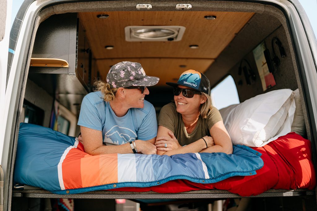 A couple laying in the back of their Sprinter van smiling at each other.