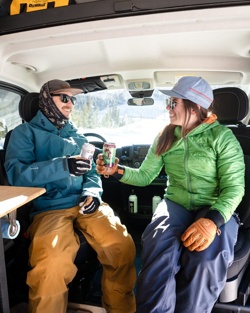 A couple clinking beers together sitting in their camper van after a day on the ski slopes.
