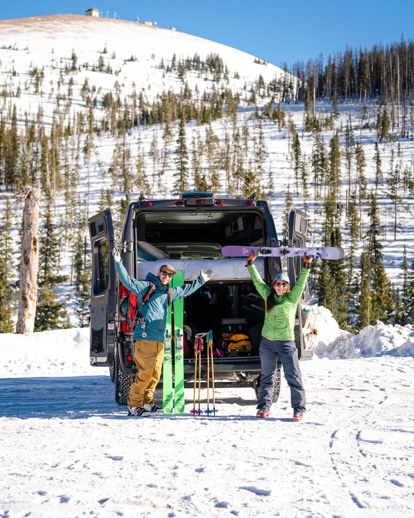 A couple standing at the back of their camper van with ski gear on excited to hit the slopes.